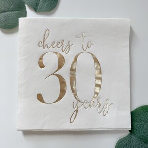 K&ouml;nnte beinhalten: Eine quadratische wei&szlig;e Papierserviette mit dem Text cheers to 30 years in metallischer Goldfolie. Die W&ouml;rter cheers to stehen in Schreibschrift &uuml;ber der gro&szlig;en Zahl 30, das Wort years steht in kleinerer Schreibschrift darunter.