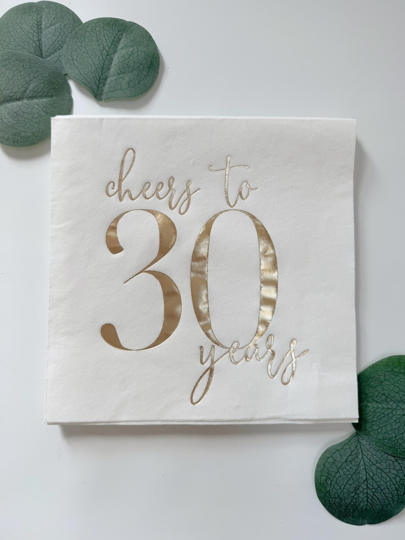 K&ouml;nnte beinhalten: Eine quadratische wei&szlig;e Papierserviette mit dem Text cheers to 30 years in metallischer Goldfolie. Die W&ouml;rter cheers to stehen in Schreibschrift &uuml;ber der gro&szlig;en Zahl 30, das Wort years steht in kleinerer Schreibschrift darunter.