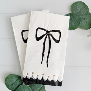 May include: Two white paper napkins with a black bow design and a scalloped border. The bow has a gold center and the border has gold dots. Green leaves are in the foreground.