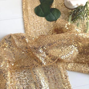 May include: Gold sequin fabric draped across a white surface. The fabric is heavily embellished with small, round sequins, creating a shimmering effect. Green leaves and a white cake stand with a cupcake are visible.