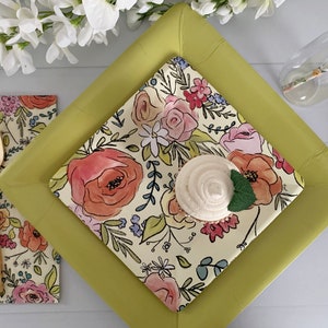 Puede incluir: Un plato cuadrado de papel amarillo con un dise&ntilde;o floral. El plato est&aacute; cubierto con un plato de papel blanco m&aacute;s peque&ntilde;o con un dise&ntilde;o floral. Un cupcake con glaseado blanco est&aacute; en el plato m&aacute;s peque&ntilde;o. Un tenedor y un cuchillo dorados est&aacute;n en la mesa.