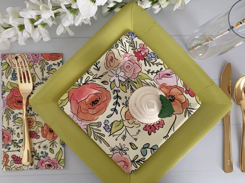 Puede incluir: Un plato cuadrado de papel amarillo con un dise&ntilde;o floral. El plato est&aacute; cubierto con un plato de papel blanco m&aacute;s peque&ntilde;o con un dise&ntilde;o floral. Un cupcake con glaseado blanco est&aacute; en el plato m&aacute;s peque&ntilde;o. Un tenedor y un cuchillo dorados est&aacute;n en la mesa.