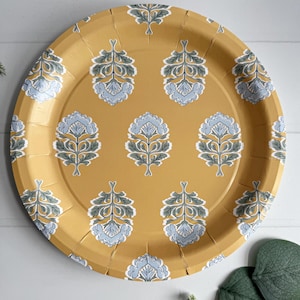 May include: Round, mustard-yellow paper plate with a repeating floral pattern in light blue, gray, and white. The plate has a slightly textured surface and is suitable for serving food at a party or event.