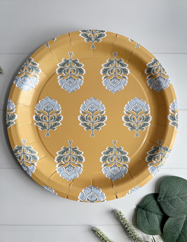 May include: Round, mustard-yellow paper plate with a repeating floral pattern in light blue, gray, and white. The plate has a slightly textured surface and is suitable for serving food at a party or event.