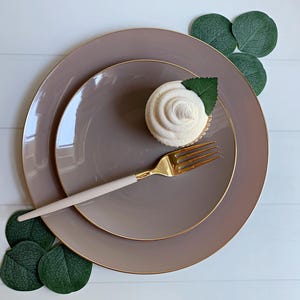 May include: A top-down shot of a table setting featuring two taupe plates with gold rims, a gold fork with a light pink handle, and a cupcake with a white swirl frosting and a green leaf. Green leaves are arranged around the plates.