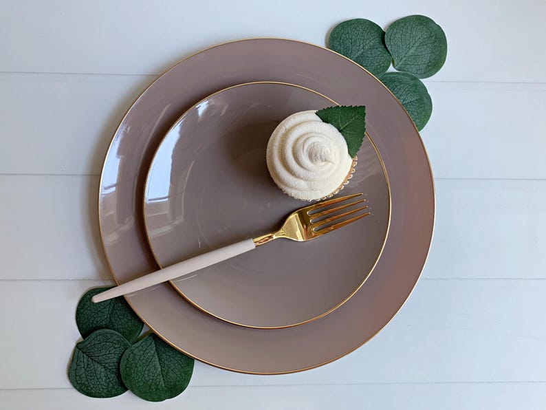 May include: A top-down shot of a table setting featuring two taupe plates with gold rims, a gold fork with a light pink handle, and a cupcake with a white swirl frosting and a green leaf. Green leaves are arranged around the plates.