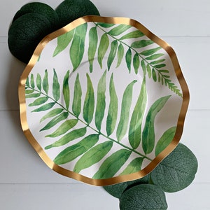 May include: A white paper party plate with a gold rim and a green fern leaf design. The plate is on a white surface with green leaves around it.