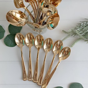 May include: A collection of gold-colored spoons, some arranged in a clear glass container, others fanned out on a white surface. The spoons have a shiny, reflective finish, and the arrangement suggests a focus on dining or serving.
