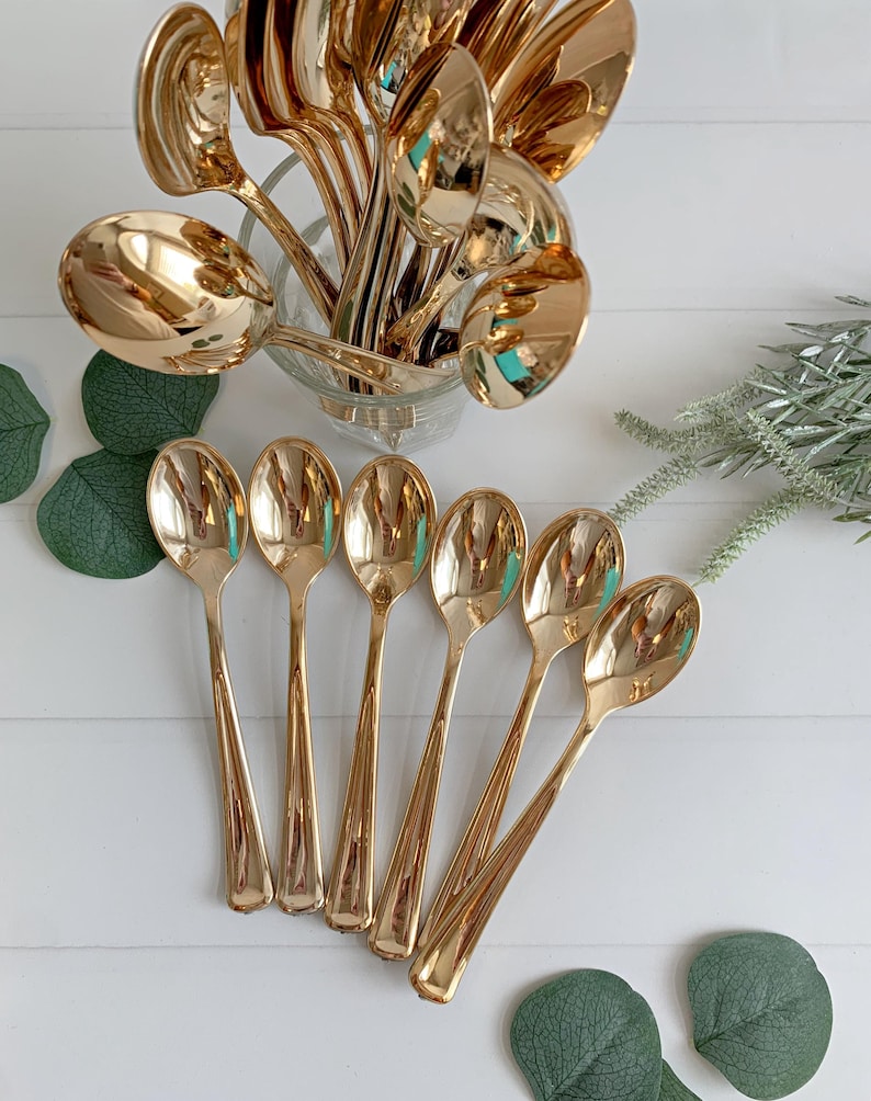May include: A collection of gold-colored spoons, some arranged in a clear glass container, others fanned out on a white surface. The spoons have a shiny, reflective finish, and the arrangement suggests a focus on dining or serving.