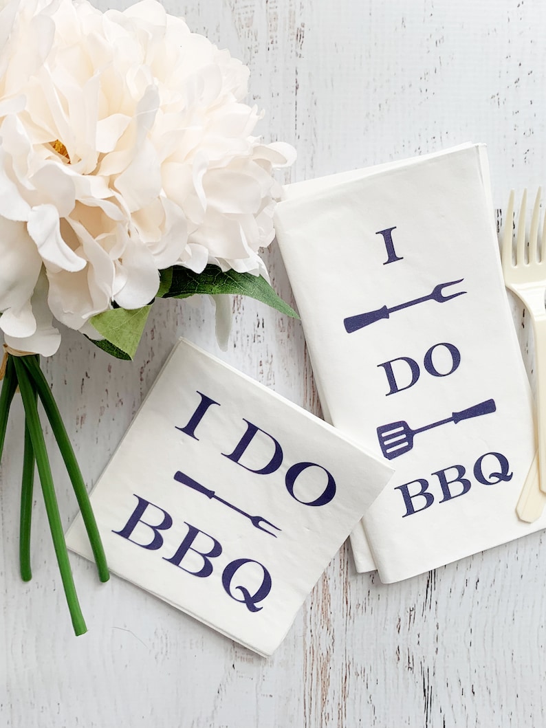 May include: Two white napkins with blue text and illustrations of grilling tools. The text on the napkins reads "I DO BBQ".