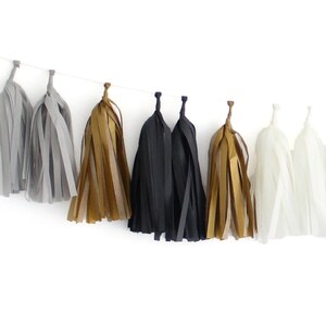 May include: A decorative garland featuring a series of hanging tassels in a repeating colour sequence of light grey, metallic gold, black, and white. Each tassel is constructed from thin, vertical strips of material suspended from a thin string.