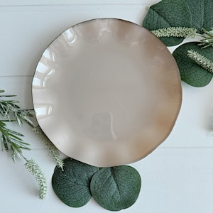 May include: A beige ceramic plate with a ruffled edge and a dark rim. The plate is surrounded by green leaves and sprigs of greenery, creating a natural, decorative arrangement. The plate's design is simple and elegant.