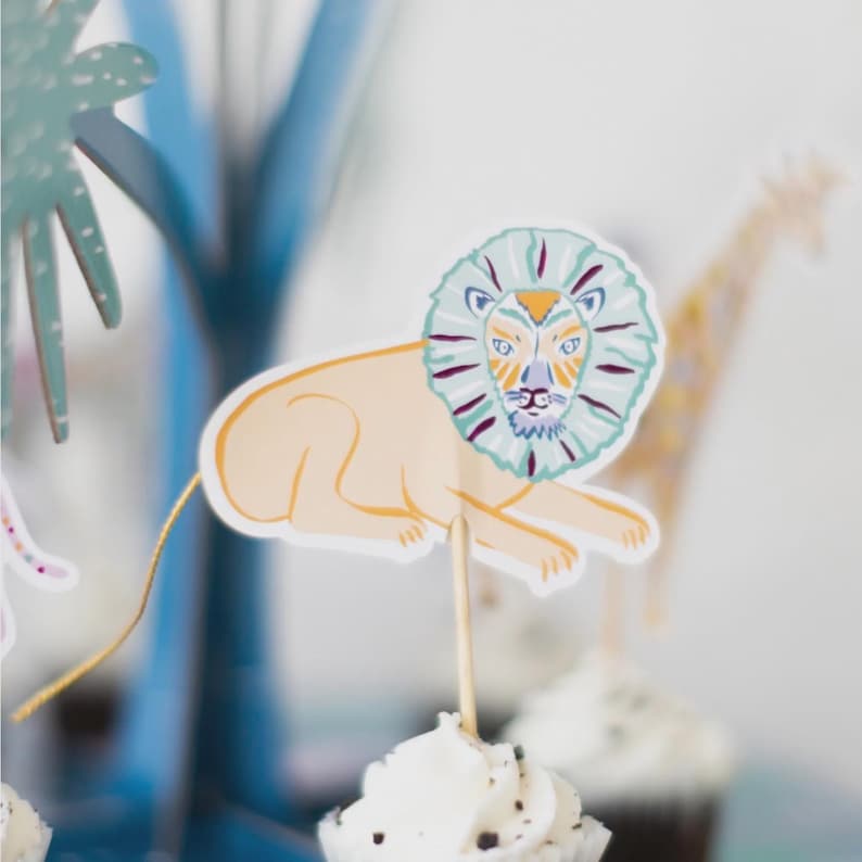 K&ouml;nnte beinhalten: Ein Cupcake mit wei&szlig;em Zuckerguss ist mit einer L&ouml;wen-Dekoration aus Papier verziert. Der L&ouml;we hat einen beigefarbenen K&ouml;rper, eine blaue und violette M&auml;hne und ein detailliertes Gesicht. Eine Giraffe und eine Palmen-Dekoration befinden sich im Hintergrund.