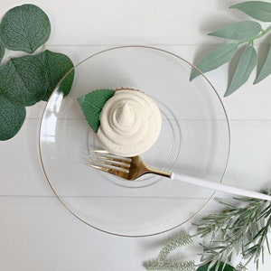 May include: A frosted cupcake with a green leaf garnish sits on a clear glass plate with a gold rim. A gold fork with a white handle rests beside the cupcake. The background features green leaves and a white wooden surface.