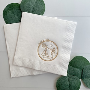 May include: Two white paper napkins with a gold foil design. The design features a tent, a moon, stars, and the text "Katie & Jake 12.02.2023".