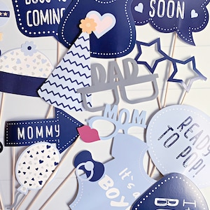 May include: A collection of baby shower photo booth props on sticks, featuring navy and light blue designs. Items include signs reading BOSS IS COMING, COMING SOON, MOMMY, READY TO POP, and IT'S A BOY, plus glasses, a party hat, and a baby bib.