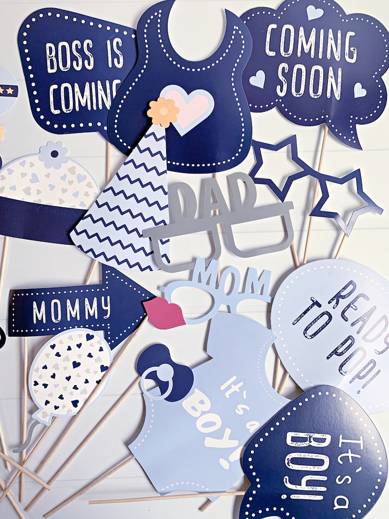 May include: A collection of baby shower photo booth props on sticks, featuring navy and light blue designs. Items include signs reading BOSS IS COMING, COMING SOON, MOMMY, READY TO POP, and IT'S A BOY, plus glasses, a party hat, and a baby bib.