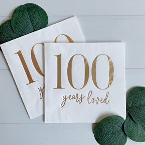 May include: Two square white paper napkins featuring the metallic gold text 100 years loved. The napkins are decorated with dark green eucalyptus leaves placed at the top left and bottom right corners of the composition.