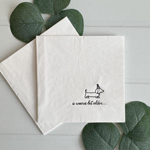 May include: A square white paper napkin featuring a black line drawing of a dachshund wearing a party hat. Below the dog, the text reads a weenie bit older... in a black cursive font. The napkin is folded and presented as a single unit.
