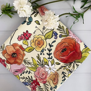 May include: A square, cream-colored decorative plate with a vibrant floral design. The plate features hand-painted flowers in shades of red, orange, pink, and yellow, with green leaves and stems. White flowers and greenery are placed on top.