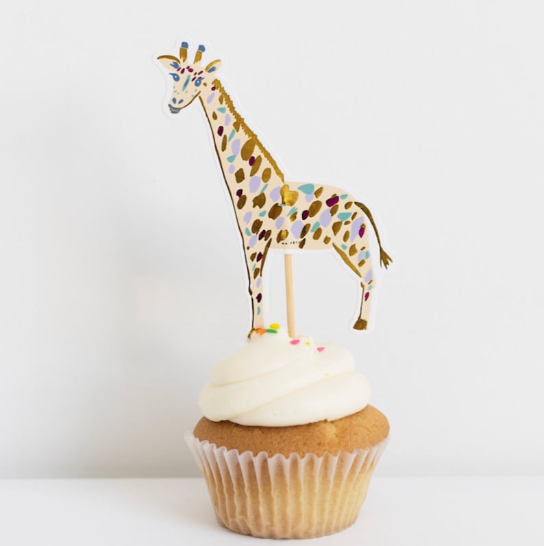 May include: A cupcake topped with white frosting and colorful sprinkles, decorated with a giraffe cupcake topper. The giraffe is illustrated in gold, purple, and blue.
