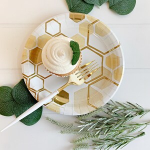 May include: A white paper plate with a gold honeycomb pattern, a cupcake with white frosting and a green mint leaf on top, and a gold fork on the plate. Green eucalyptus leaves and sprigs of greenery are scattered around the plate.