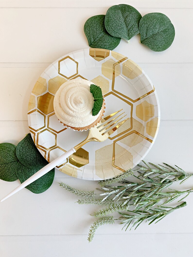 May include: A white paper plate with a gold honeycomb pattern, a cupcake with white frosting and a green mint leaf on top, and a gold fork on the plate. Green eucalyptus leaves and sprigs of greenery are scattered around the plate.