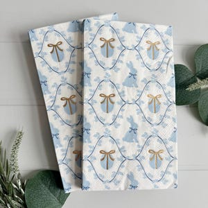 May include: Two white paper napkins with a blue and gold pattern. The pattern features a repeating design of bunnies, leaves, and eggs with gold bows.