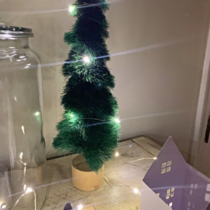 May include: A small green artificial Christmas tree with white fairy lights on a wooden table with a white house shaped decoration and a clear glass jar.