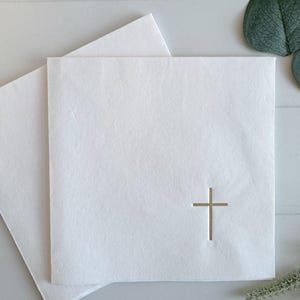 May include: A square white paper napkin featuring a small, centered gold foil cross emblem on the lower right quadrant. The napkin is folded and layered, displaying a smooth, matte texture with clean edges and a minimalist design.