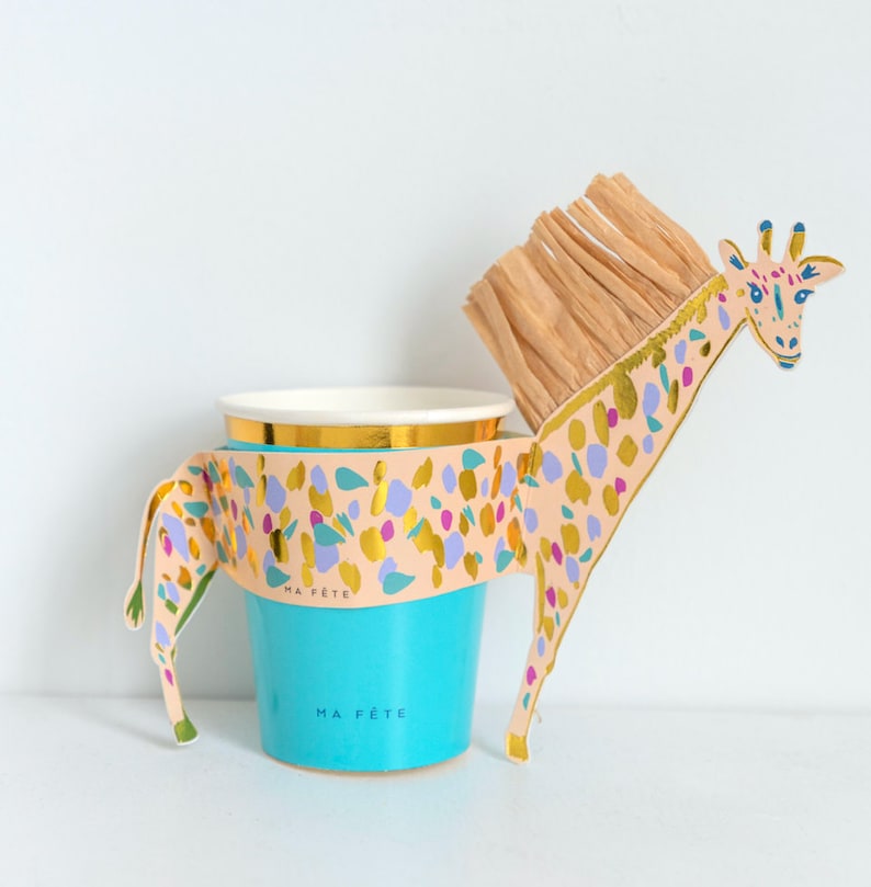 K&ouml;nnte beinhalten: Ein t&uuml;rkisfarbener Pappbecher mit goldenem Rand und einem halter in Form einer Giraffe. Der Halter ist pfirsichfarben mit goldenen, lila und blauen Konfetti-&auml;hnlichen Designs. Die Giraffe hat eine braune M&auml;hne und die Worte "MA F&Ecirc;TE" sind auf dem Becher aufgedruckt.