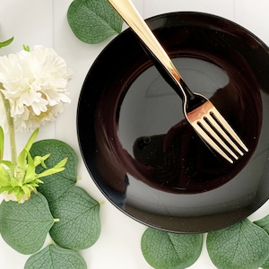 May include: A black plate with a gold fork on top. The plate is surrounded by green leaves.