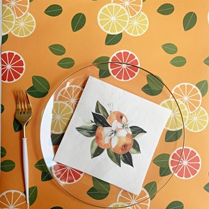 Op de afbeelding: Een heldere glazen plaat en een gouden vork liggen op een levendig oranje tafelkleed met een patroon van citrusvruchtenschijfjes en groene bladeren. Een vierkante servet met een aquarelillustratie van sinaasappels en bloesems ligt op de plaat.