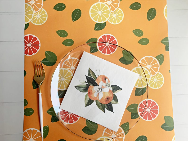 Op de afbeelding: Een heldere glazen plaat en een gouden vork liggen op een levendig oranje tafelkleed met een patroon van citrusvruchtenschijfjes en groene bladeren. Een vierkante servet met een aquarelillustratie van sinaasappels en bloesems ligt op de plaat.