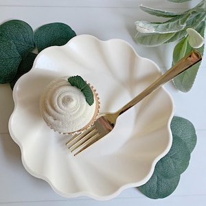 May include: A white cupcake with white frosting and a mint leaf sits on a scalloped white plate. A gold fork rests on the plate. Green leaves and a white background complete the scene, suggesting a dessert or food-related theme.