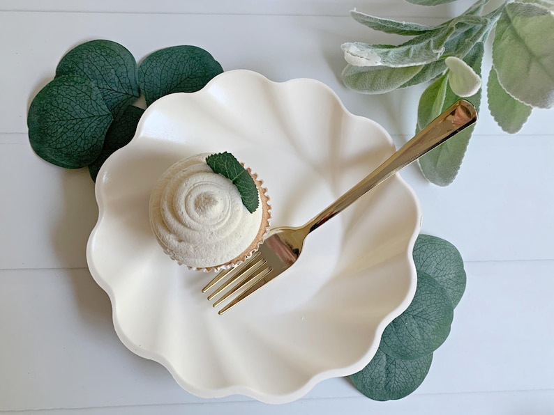 May include: A white cupcake with white frosting and a mint leaf sits on a scalloped white plate. A gold fork rests on the plate. Green leaves and a white background complete the scene, suggesting a dessert or food-related theme.