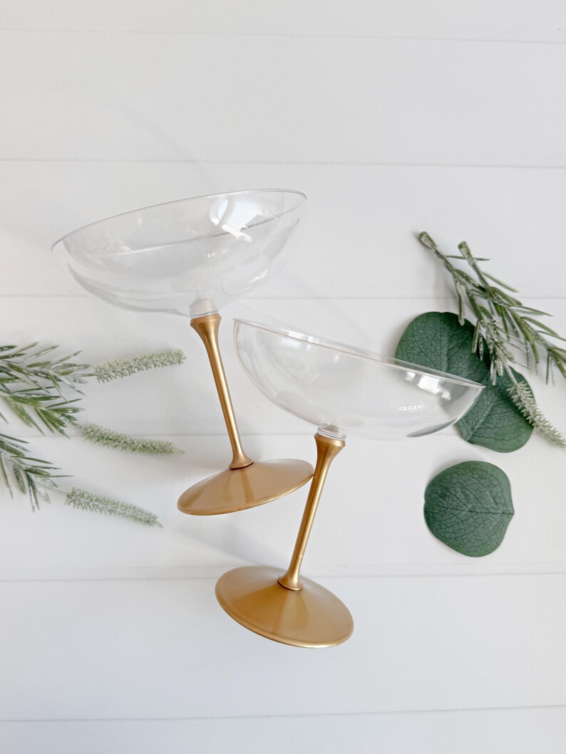 Puede incluir: Dos copas de champ&aacute;n transparentes con tallos y bases doradas. Las copas est&aacute;n colocadas sobre una superficie blanca, con acentos de follaje verde. Las copas tienen un dise&ntilde;o de cuenco ancho y poco profundo.