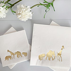 May include: Two stacks of white napkins with gold foil animal silhouettes. The top napkin in each stack features a line of animals including a rhinoceros, giraffe, elephant, and lion.