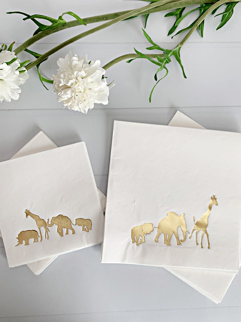 May include: Two stacks of white napkins with gold foil animal silhouettes. The top napkin in each stack features a line of animals including a rhinoceros, giraffe, elephant, and lion.