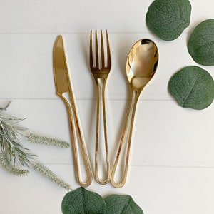 May include: Gold-colored flatware set including a knife, fork, and spoon. The utensils are arranged on a white surface, accompanied by green eucalyptus leaves and sprigs. The set is designed for elegant dining.