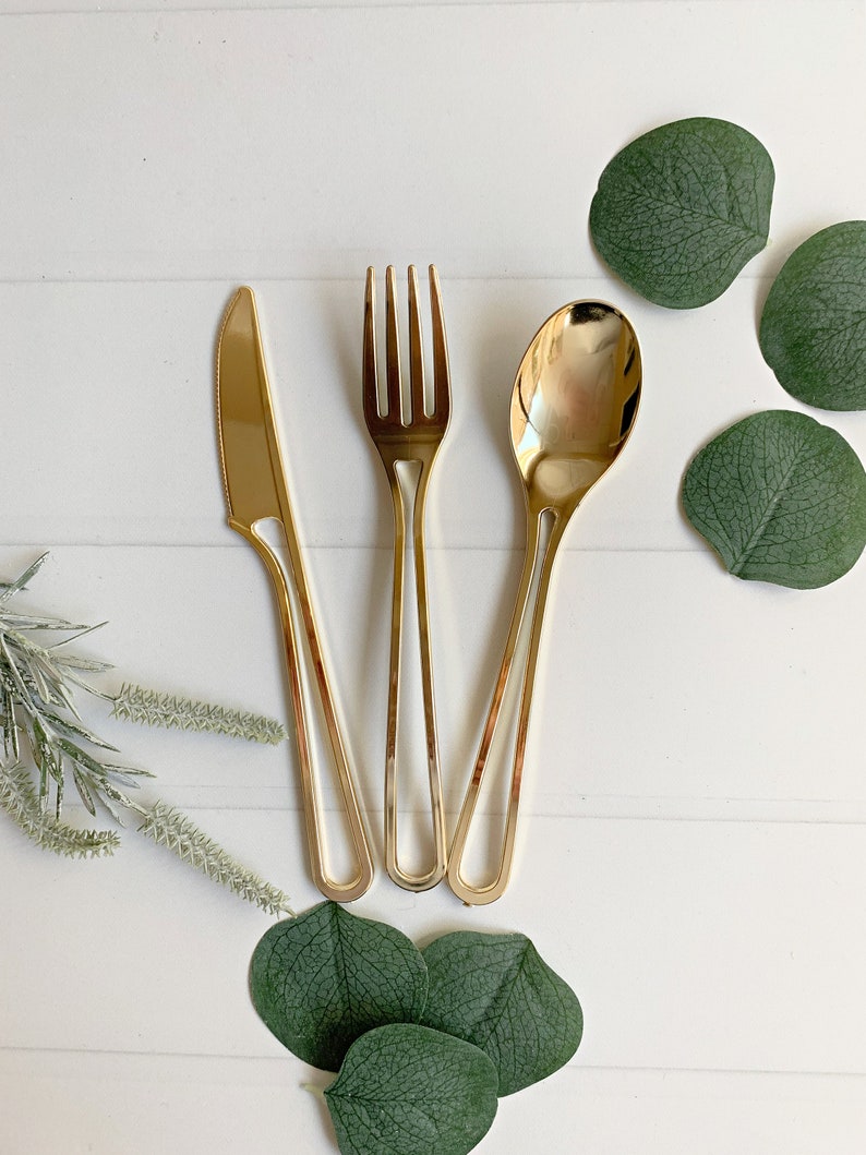 May include: Gold-colored flatware set including a knife, fork, and spoon. The utensils are arranged on a white surface, accompanied by green eucalyptus leaves and sprigs. The set is designed for elegant dining.