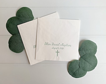 May include: Two white napkins with green text that reads "Ethan David's Baptism August 5, 2023" and a green cross. The napkins are on a white wooden surface with green eucalyptus leaves.