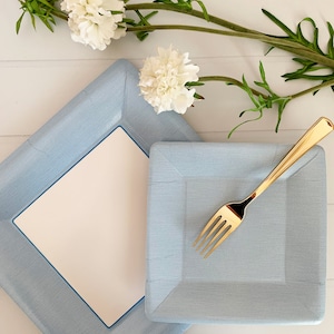 May include: Two light blue square paper plates with a white center and a gold fork on top of the smaller plate. The plates are on a white wooden surface with white flowers and green stems.