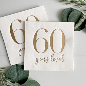 May include: Two square white paper napkins featuring the number 60 printed in a large metallic gold font. Below the number, the text years loved is written in a smaller, elegant gold cursive script. The napkins have a smooth finish.