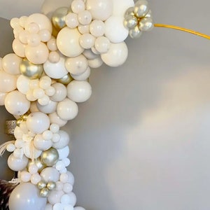 Puede incluir: Un arco de globos en tonos blancos, crema y dorados. El arreglo presenta globos de varios tama&ntilde;os, creando un efecto texturizado y en capas. El arco est&aacute; sostenido por una estructura met&aacute;lica dorada, ideal para una fiesta.