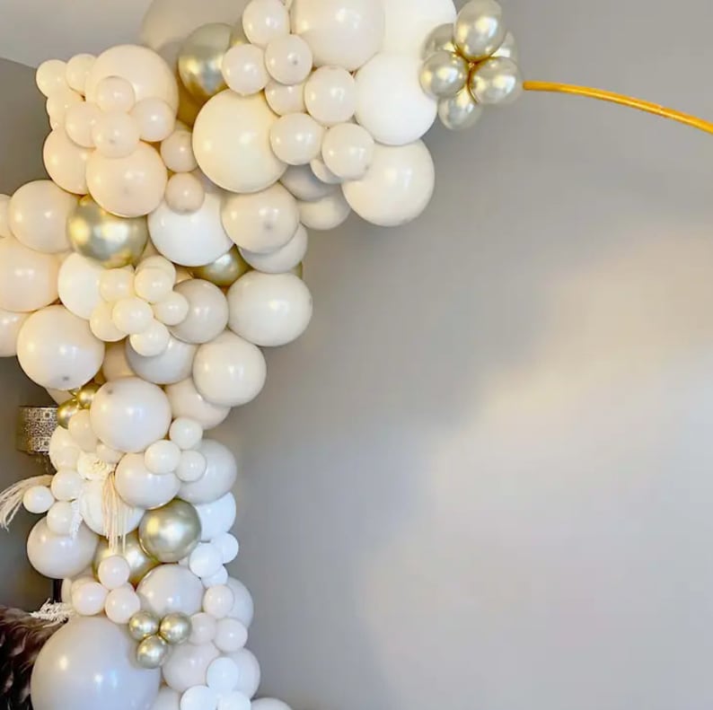 Puede incluir: Un arco de globos en tonos blancos, crema y dorados. El arreglo presenta globos de varios tama&ntilde;os, creando un efecto texturizado y en capas. El arco est&aacute; sostenido por una estructura met&aacute;lica dorada, ideal para una fiesta.