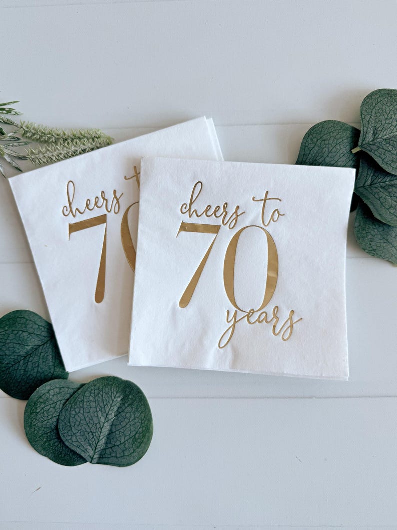 May include: Two white paper napkins with gold foil lettering. The top napkin says "Cheers to 70 years". The bottom napkin says "Cheers to 70".