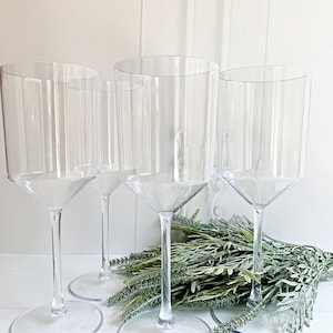 Puede incluir: Tres copas de vino de pl&aacute;stico transparente con tallo y base ancha. Las copas est&aacute;n dispuestas en fila sobre una superficie blanca con follaje verde en el centro.