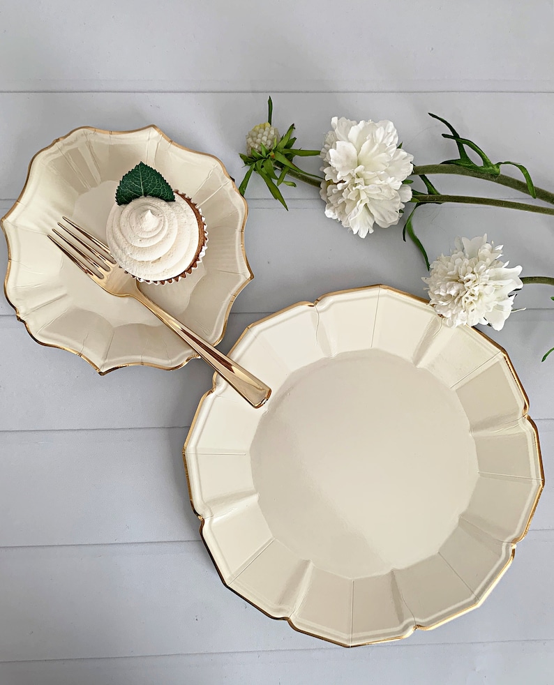 Op de afbeelding: Ivoor papieren borden met geschulpte randen en gouden rand. Een cupcake met witte glazuur en een muntblad ligt op een kleiner bord, met een gouden vork. Witte bloemen en groen op de achtergrond.