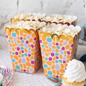 May include: Three popcorn containers with a colorful geometric pattern and gold trim. The containers are filled with popcorn and are sitting on a white surface.